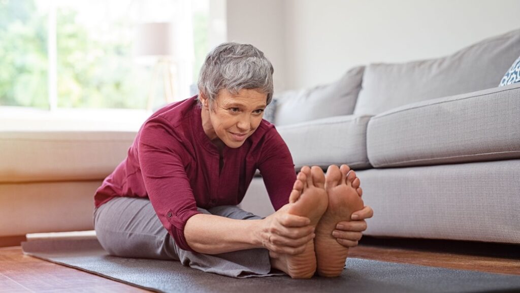 A woman sitting on the ground on a yoga mat in front of a couch bending forward and holding onto her feet with both hands